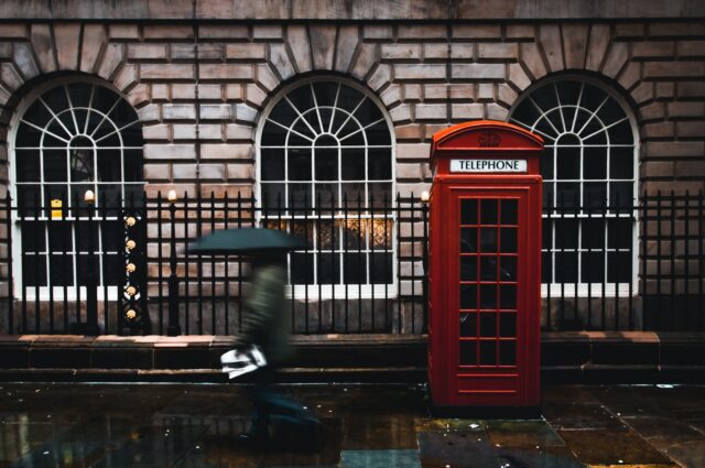 Foto af Jack Finnigan time lapse fotografering af kvinde, der går på gaden, mens hun holder paraply i nærheden af London telefonboks ved siden af væggen